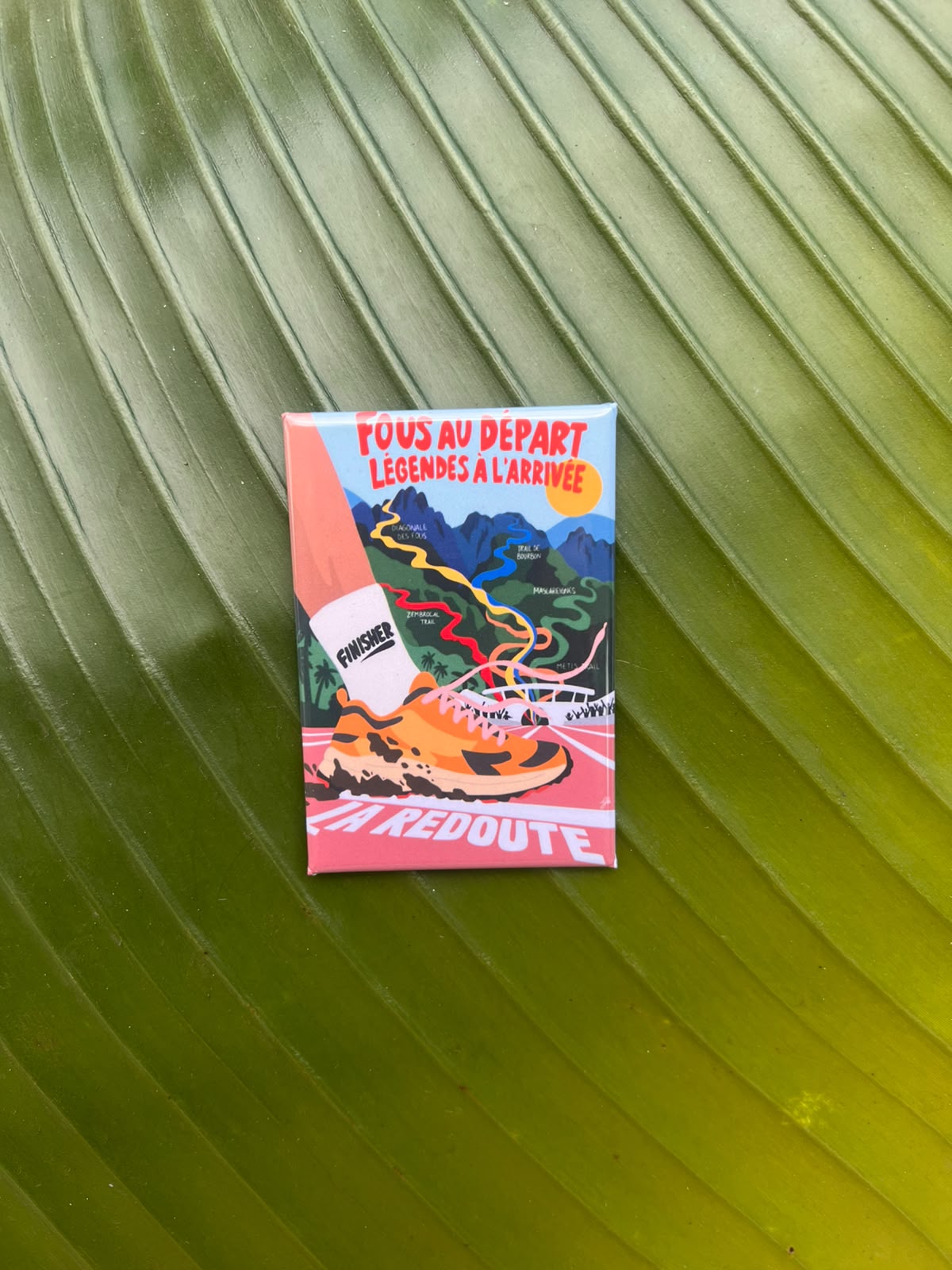 Magnet souvenir du Grand Raid de La Réunion illustré par Lefilariane, représentant des chaussures de trail sur un sentier de montagne et le message « Force à vous bande de fous », photographié sur une feuille tropicale verte.