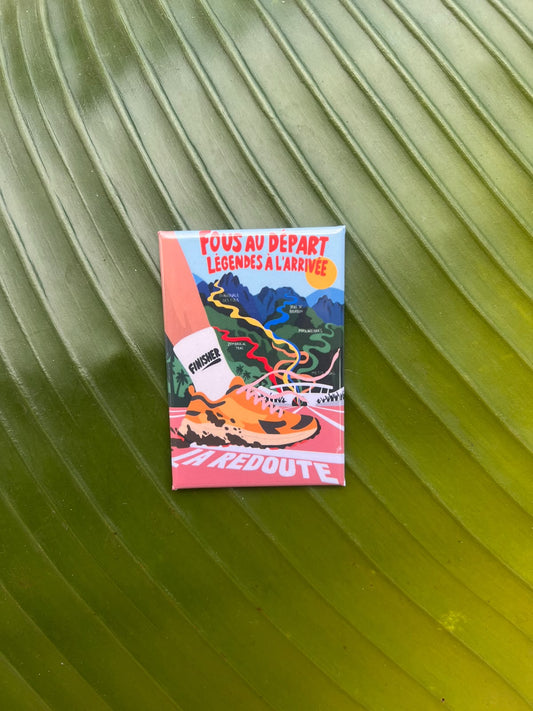 Magnet souvenir du Grand Raid de La Réunion illustré par Lefilariane, représentant des chaussures de trail sur un sentier de montagne et le message « Force à vous bande de fous », photographié sur une feuille tropicale verte.
