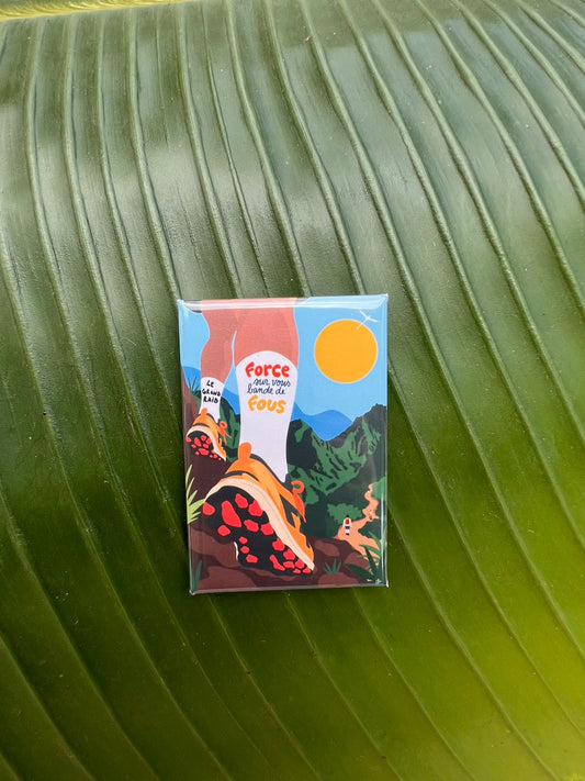 Magnet souvenir du Grand Raid de La Réunion illustré par Lefilariane, représentant des chaussures de trail sur un sentier de montagne et le message « Force à vous bande de fous », photographié sur une feuille tropicale verte. 